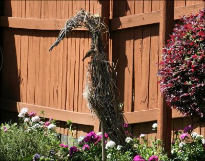 Garden sculpture of a heron, made of vines and grasses. A sparrow borrows some material for its nest.